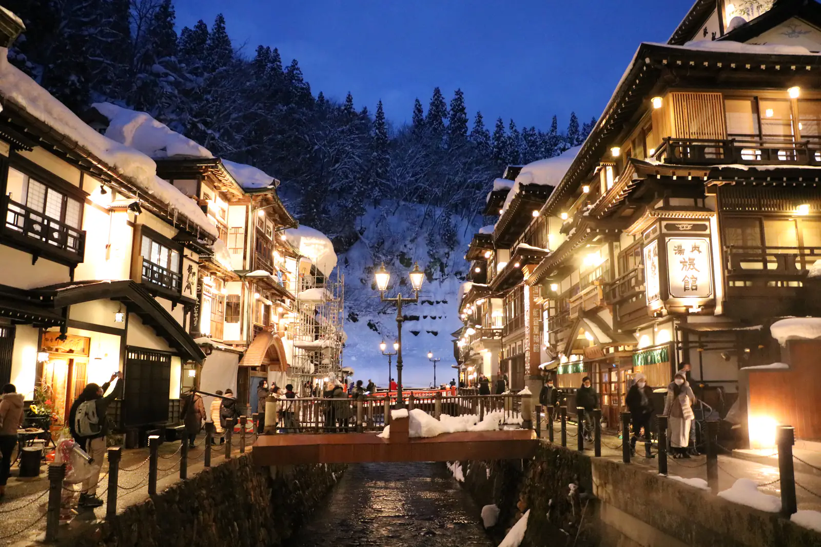 A snowy onsen town at blue hour: multi-storey wooden inns glow with warm light on both sides of a river, gas lamps lit on the bridge