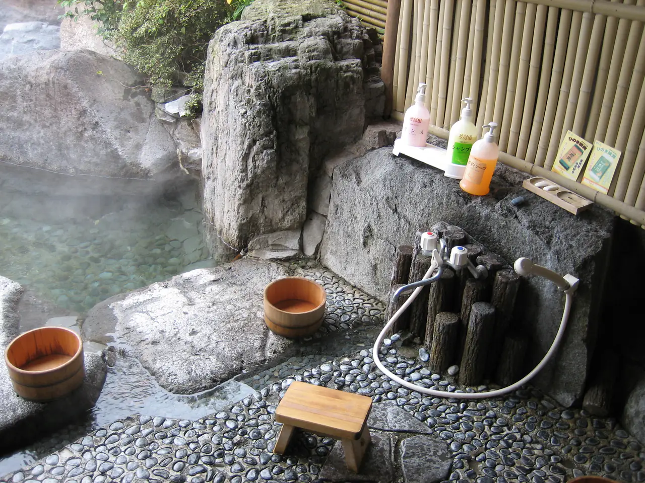 A stone washing area beside an outdoor bath, with wooden buckets, a small wooden stool and a hot-water spout