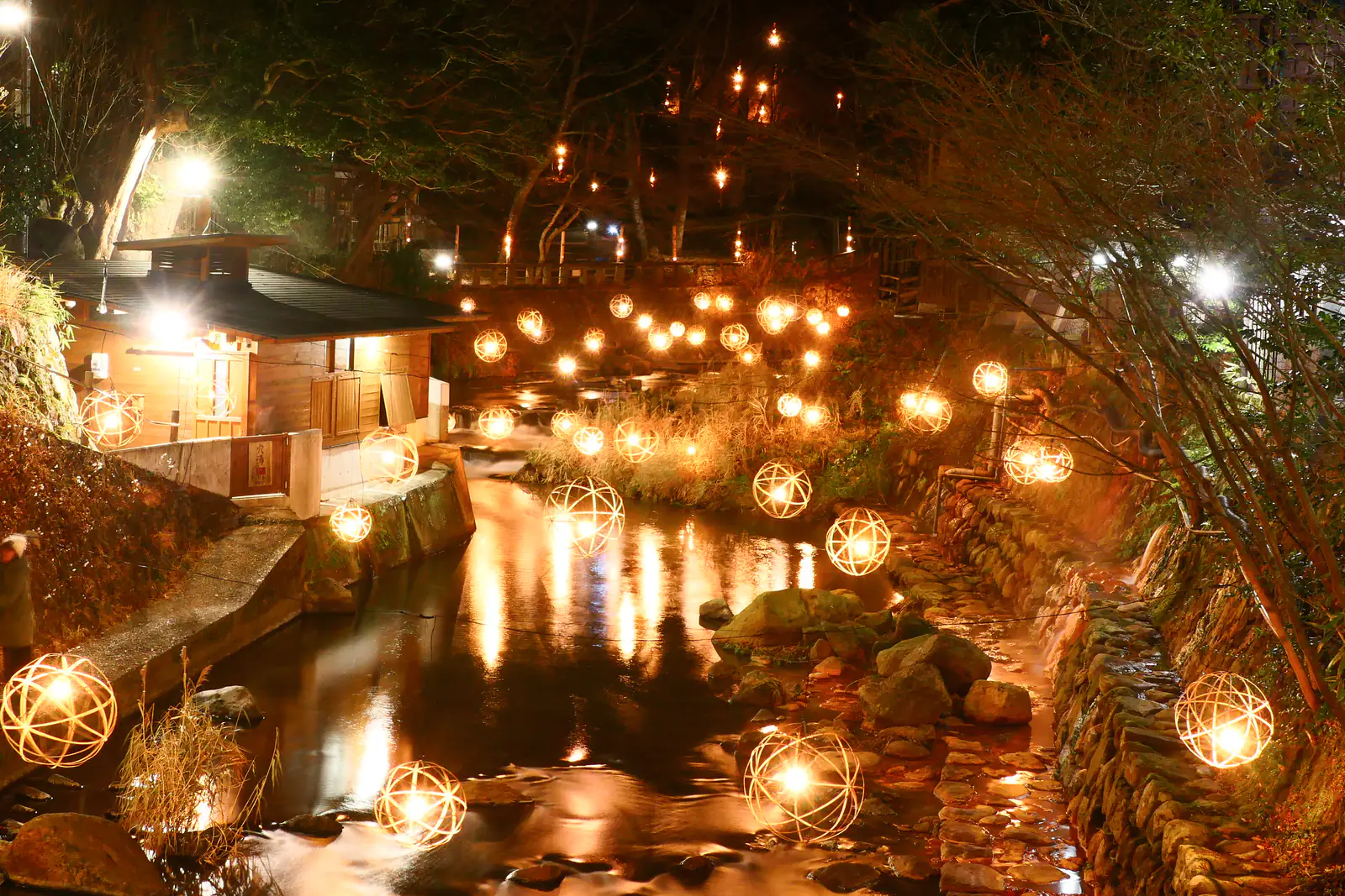 An onsen town at night: dozens of glowing round lanterns line a quiet stream beneath trees, warm light reflecting in the water