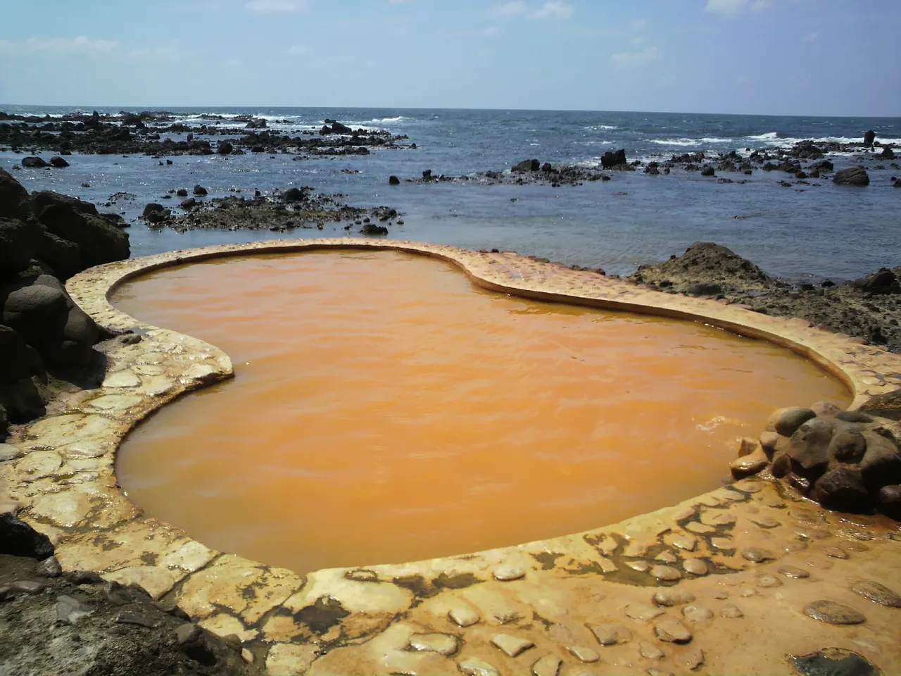 A gourd-shaped pool of rust-orange spring water on the rocky shore, the open sea just beyond