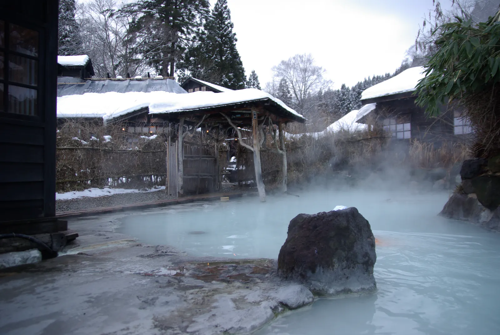 A steaming milky-white outdoor bath with a single large rock, surrounded by snow-laden roofs and winter trees