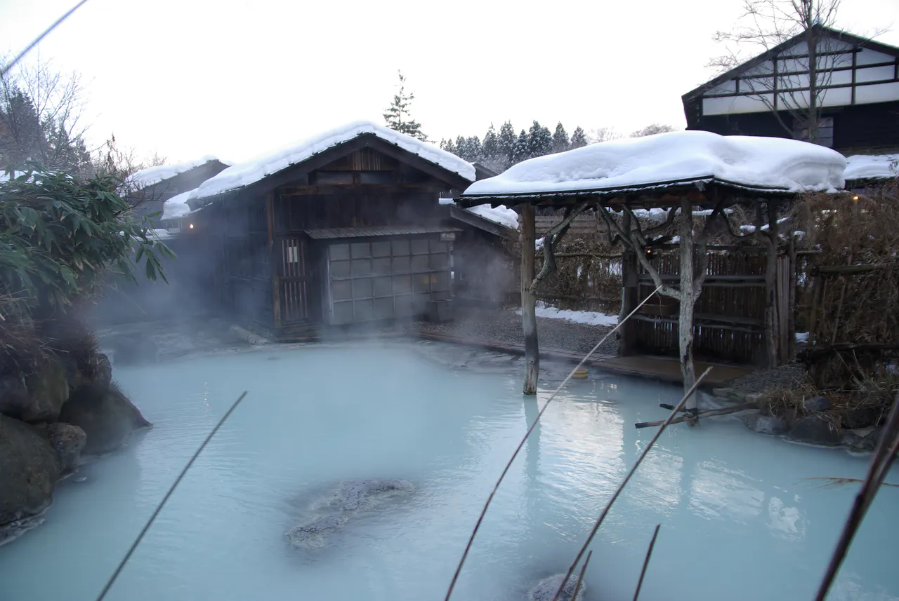 A milky-blue outdoor bath steaming in deep snow, surrounded by snow-covered wooden bathhouses