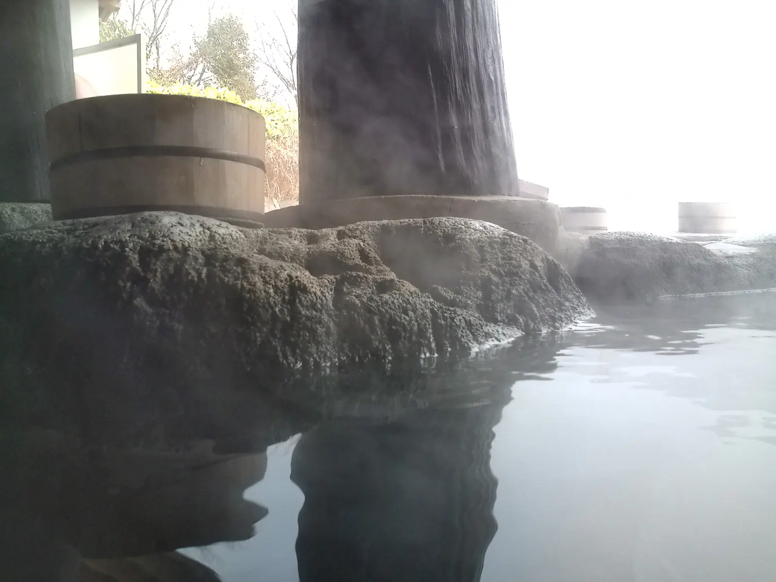 Thick white steam hanging over the still surface of an outdoor bath, a wooden bucket resting on the stone edge