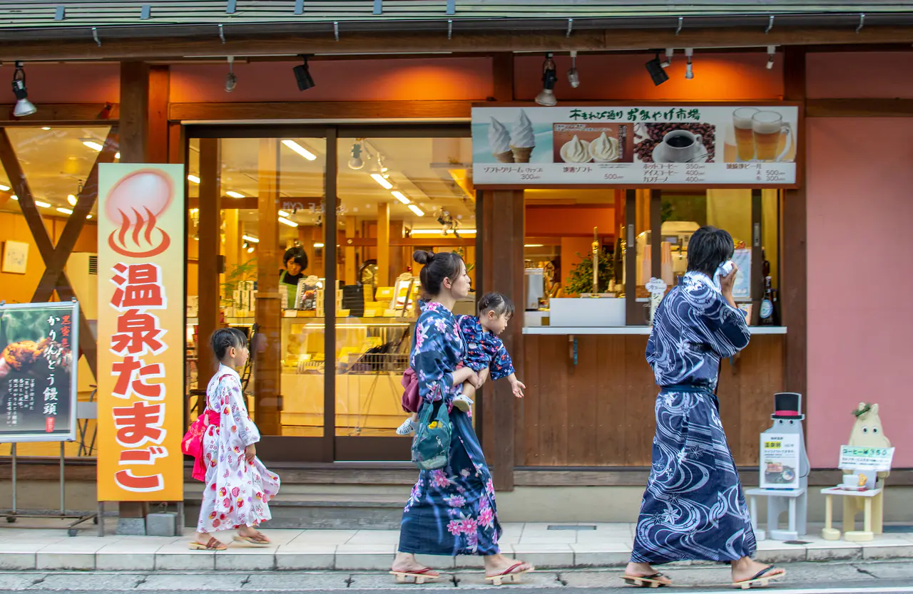 Two people in indigo yukata and a child in a pink yukata walk past a shop selling onsen eggs on an onsen-town street
