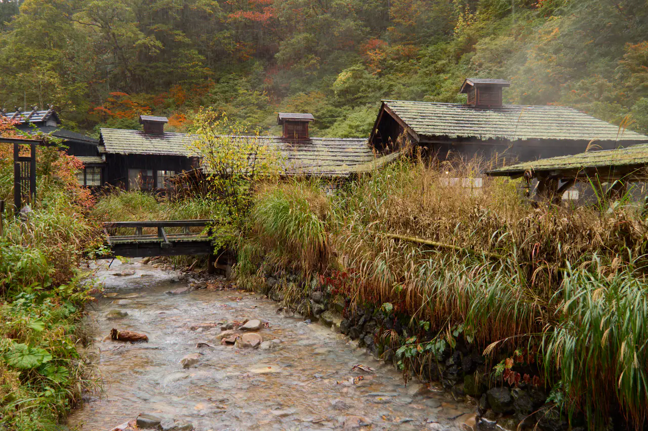 Steam rising from a rocky stream between old wooden onsen buildings, the hillside behind glowing with autumn color