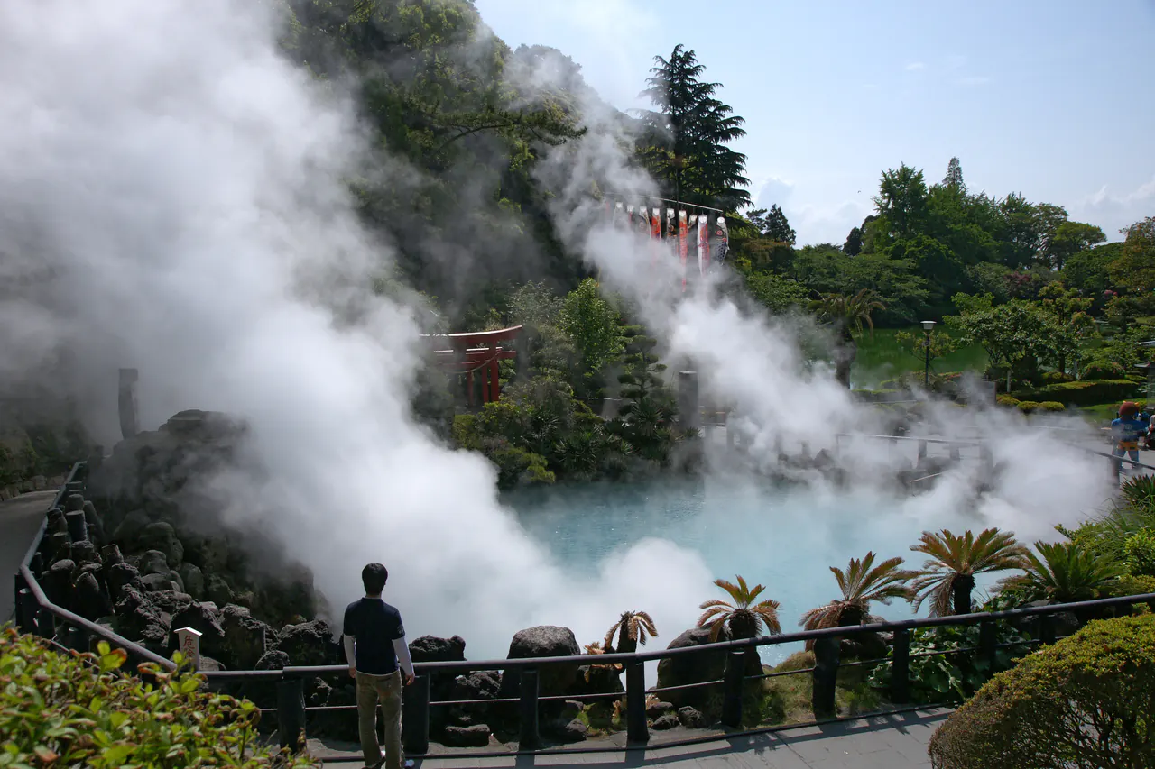 Billowing white steam rising from a vivid cobalt-blue hot spring pond surrounded by greenery, a visitor standing at the railing