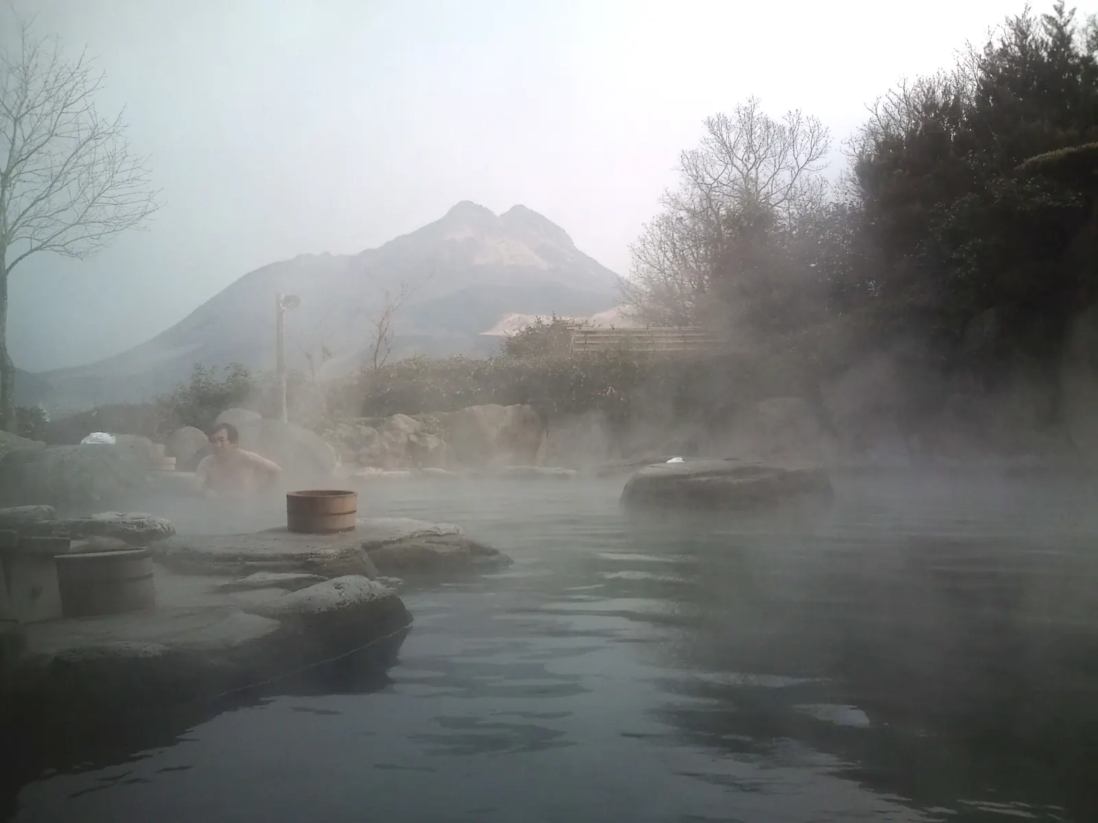 A bather soaks in a misty outdoor stone bath at dawn, steam drifting across the water and the twin peaks of Mount Yufu rising behind bare trees