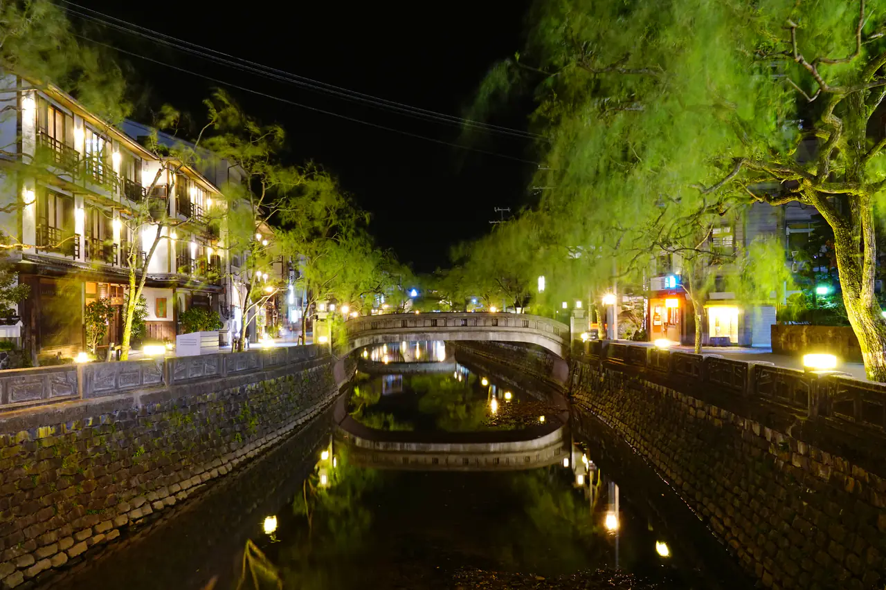 A willow-lined canal at night in Kinosaki, with a stone arched bridge and softly lit wooden buildings reflected in the water
