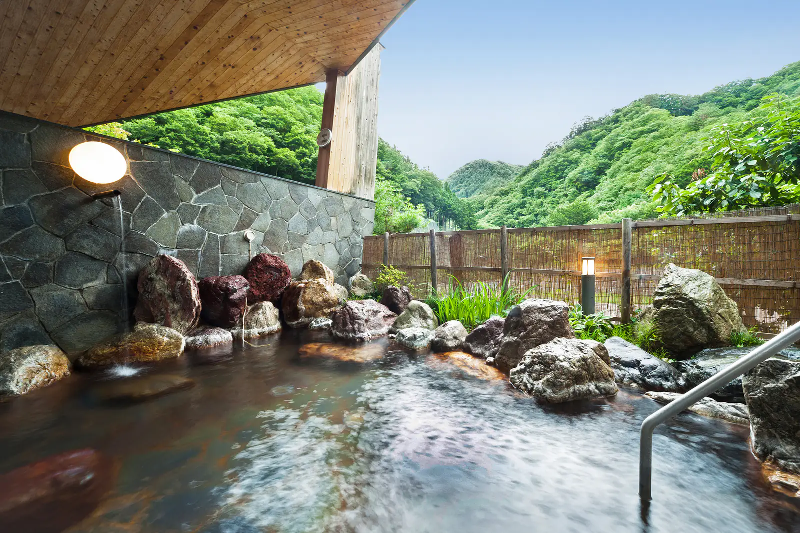 An outdoor bath of clear water among dark rocks under a wooden eave, facing lush green summer mountains