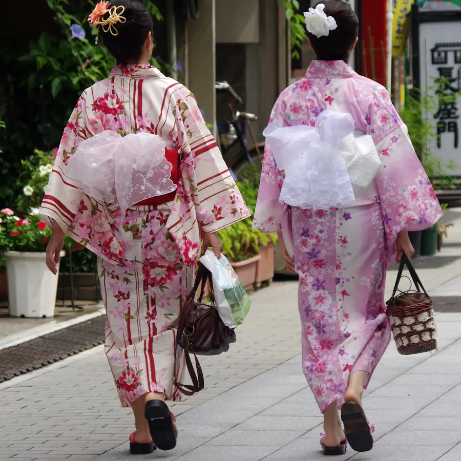 Two women in pink floral yukata walk away down a street, with wide obi tied in bows at their backs