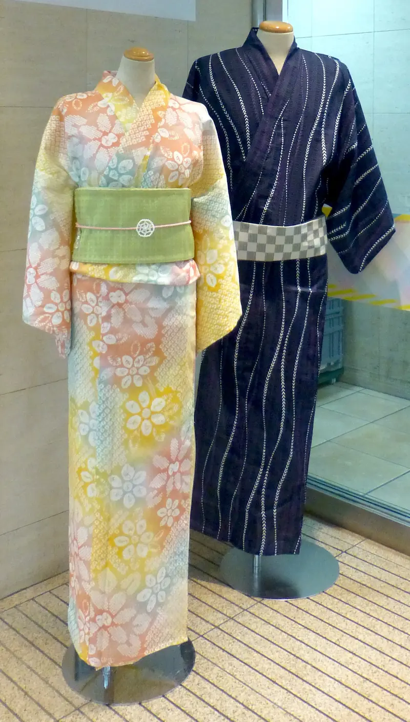 A women’s yukata with a wide green obi at the waist beside a men’s navy yukata with a narrow checked obi worn lower, on display stands