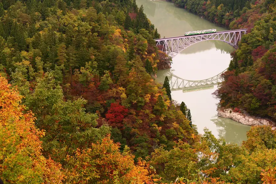 An iron arch bridge of the Tadami Line crossing the green Tadami River between red and orange autumn mountains