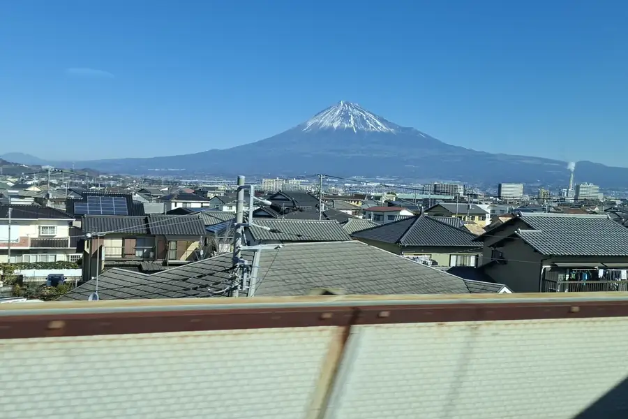 Mount Fuji seen through a Shinkansen window above rooftops on a clear blue day